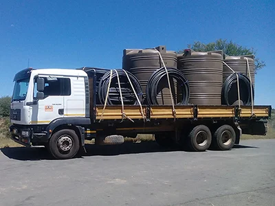 A KG Project, Plant Hire & Maintenance flatbed truck transporting heavy infrastructure components, including large industrial water tanks and coils of high-density piping, to a construction site.